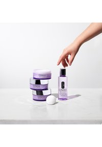 Produits de soin Clinique dans des pots violets avec des couvercles argentés, à côté d'une bouteille transparente ; un coton repose sur une surface claire.