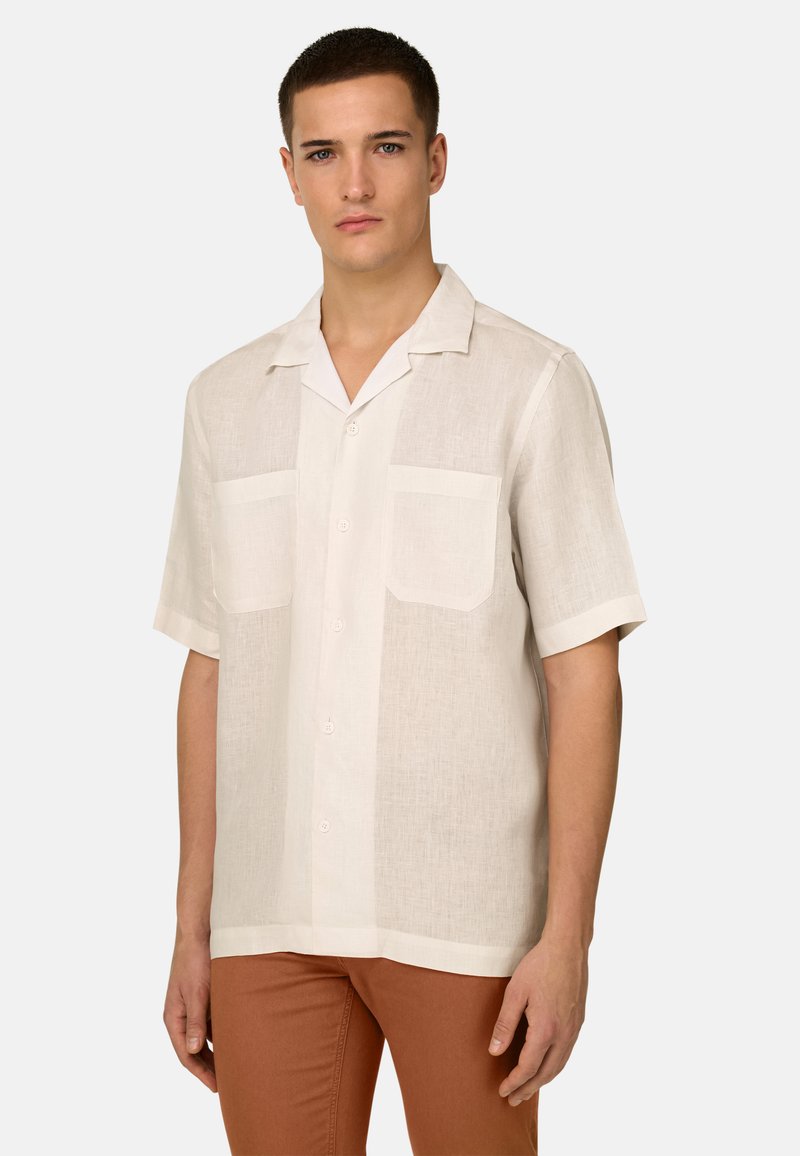 Jeune homme portant une chemise en lin blanche cassée à manches courtes, boutonnée, avec deux poches poitrine et un pantalon marron, debout les mains détendues.