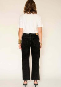 Jean noir évasé avec ourlets effilochés, en denim, associé à un t-shirt blanc à manches courtes. Bracelet au poignet, fond neutre.