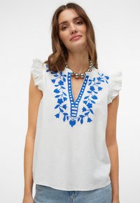 Witte mouwloze top met gerimpelde schouders, voorzien van blauwe bloemenborduursels en een V-halslijn benadrukt met verticale strepen.