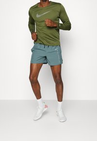 Mann in olivgrünem, langärmligem Nike-Shirt und türkisfarbenen Nike-Laufsorten, mitten im Schritt in einer Jogging- oder Laufbewegung vor weißem Hintergrund.