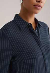 Tmavomodrá košeľa s tenkými zvislými bielymi pruhmi, zapínaný golier a krátke rukávy, vyrobená z hladkej látky.