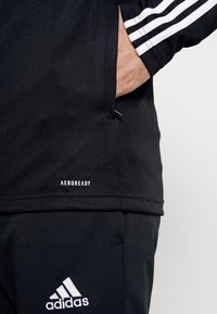 Zwarte sporttop met witte strepen op de mouwen, voorzien van een zak en "AEROREADY" tekst. Materiaal is glad en lichtgewicht.