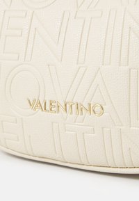 Krém színű, texturált bőrfelület, amelyen a "VALENTINO" szöveg domborított mintázatban ismétlődik, a középpontban pedig egy kis arany fém "VALENTINO" logó található.