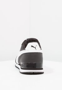Zapatilla deportiva en blanco y negro con parte superior de cuero liso, cuello acolchado y suela texturizada. Incluye un logo negro en el talón.