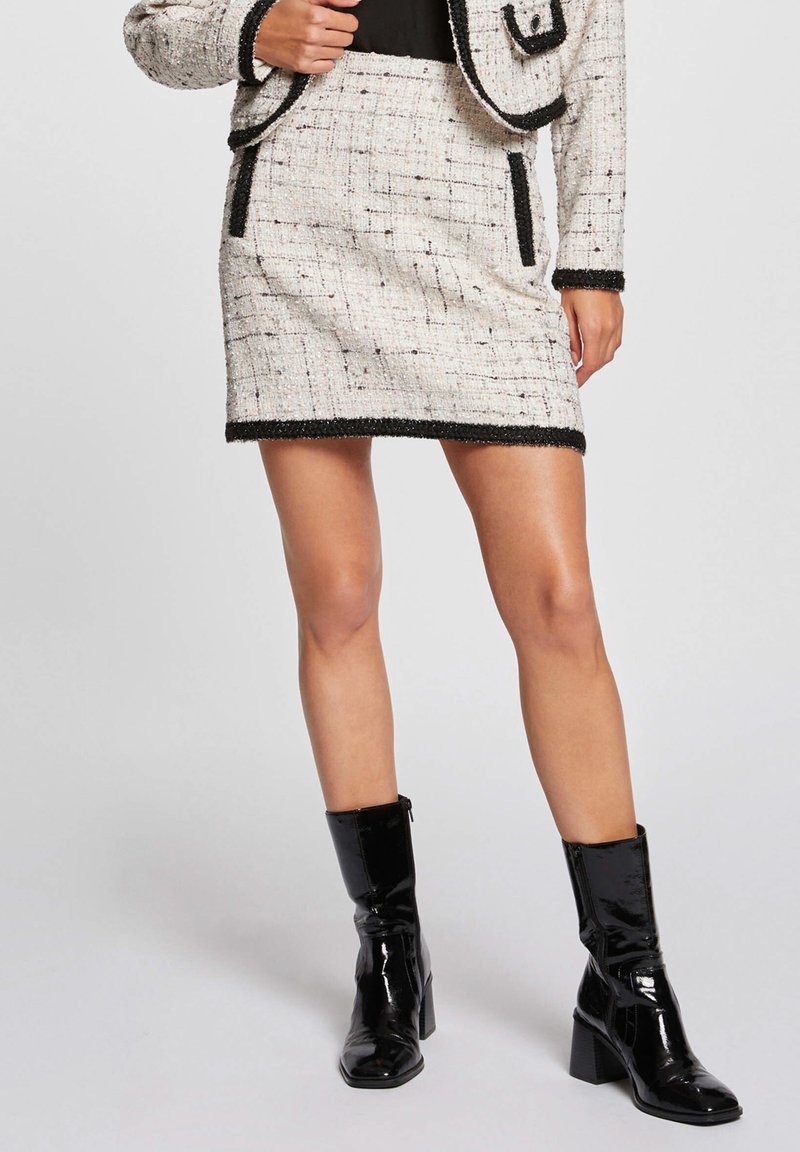 Mini falda de tweed blanca con acentos negros, acabado texturizado, que incluye dos bolsillos laterales y un dobladillo en contraste, combinada con botas de tobillo negras brillantes.