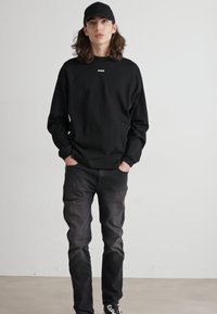 Black sweatshirt with ribbed cuffs and a small logo on the chest, paired with dark jeans and black sneakers, worn with a matching cap.