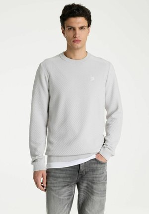Junger Mann mit lockigem dunklem Haar, der einen hellgrauen texturierten Pullover und ausgewaschene graue Jeans vor einem einfarbigen weißen Hintergrund trägt.