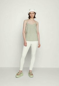 Top de alças em padrão de gingham verde e branco, calças brancas justas e sandálias de plataforma verdes. Modelo usa um chapéu balde branco. Visual simples e casual.