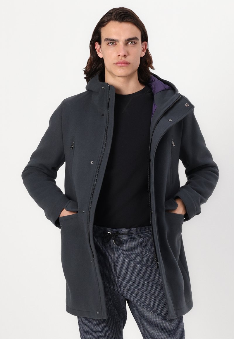 Cappotto grigio con cappuccio, dotato di cerniera frontale, due tasche laterali e una fodera viola a contrasto. Il tessuto sembra morbido e strutturato.