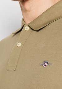 Gros plan sur un polo beige à deux boutons avec un petit logo brodé en forme de bouclier orné d'étoiles et de rayures sur la poitrine.