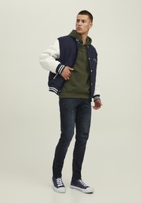 Marineblaues und weißes College-Jackett mit Kapuzenpullover in Grau, dunkle Jeans mit Dégradé-Effekten und marineblaue Sneakers. Neutraler Hintergrund.