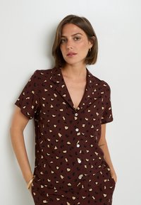 Chemise à manches courtes, de couleur marron, ornée d'un imprimé léopard en noir et crème, avec une fermeture à boutons et un col à revers.