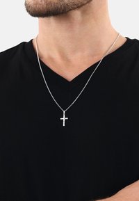 Silverkorshalsband på en delikat kedja, bärs över en svart V-ringad skjorta, med en texturerad korsdesign.