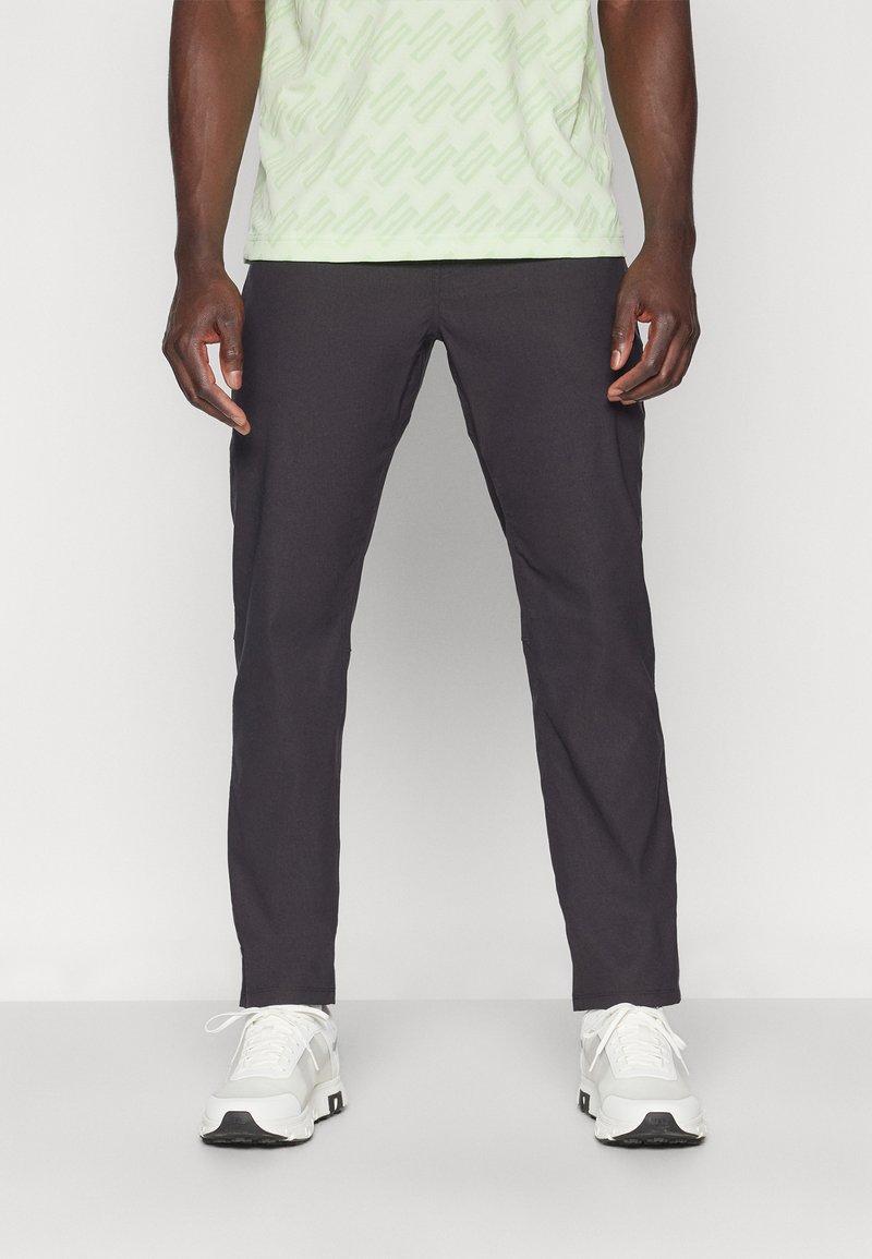 Pantalons noirs ajustés avec une texture lisse, associés à une chemise à motif vert clair et des baskets blanches avec des accents noirs.