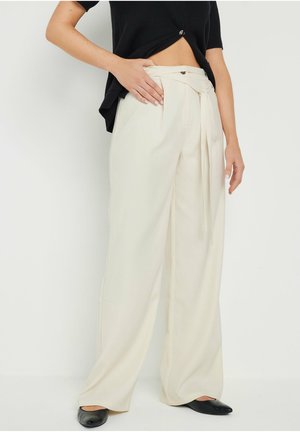 Femme portant un pantalon crème taille haute à jambes larges avec une ceinture en tissu et des ballerines noires, debout avec une main sur la hanche.