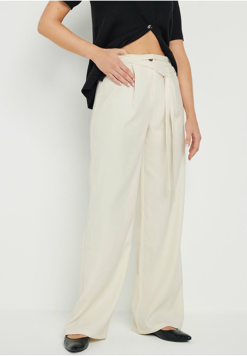 Femme portant un pantalon crème taille haute à jambes larges avec une ceinture en tissu et des ballerines noires, debout avec une main sur la hanche.
