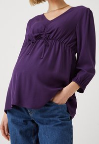 Lila gravidblus med V-ringning, samlad frontdetalj och trekvartsärmar. Mjuk tyg faller över en rundad form.