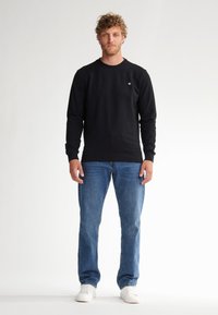 Sudadera negra de cuello redondo con puños y dobladillo de canalé, combinada con jeans de mezclilla azul y zapatillas blancas. El modelo está de pie frente a un fondo sencillo.
