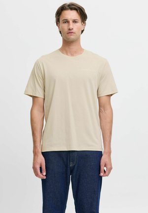 Hombre con camiseta beige de manga corta y vaqueros azul oscuro, de pie frente a un fondo blanco liso.