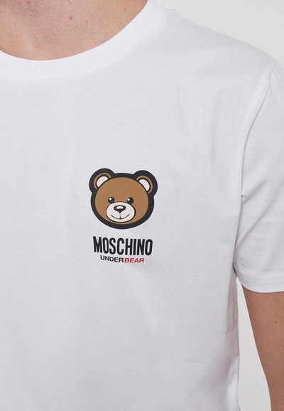 T-shirt in cotone bianco con grafica di un orso bruno e testo nero e rosso "Moschino UNDERBEAR" sotto il design dell'orso. Maniche corte e collo rotondo.