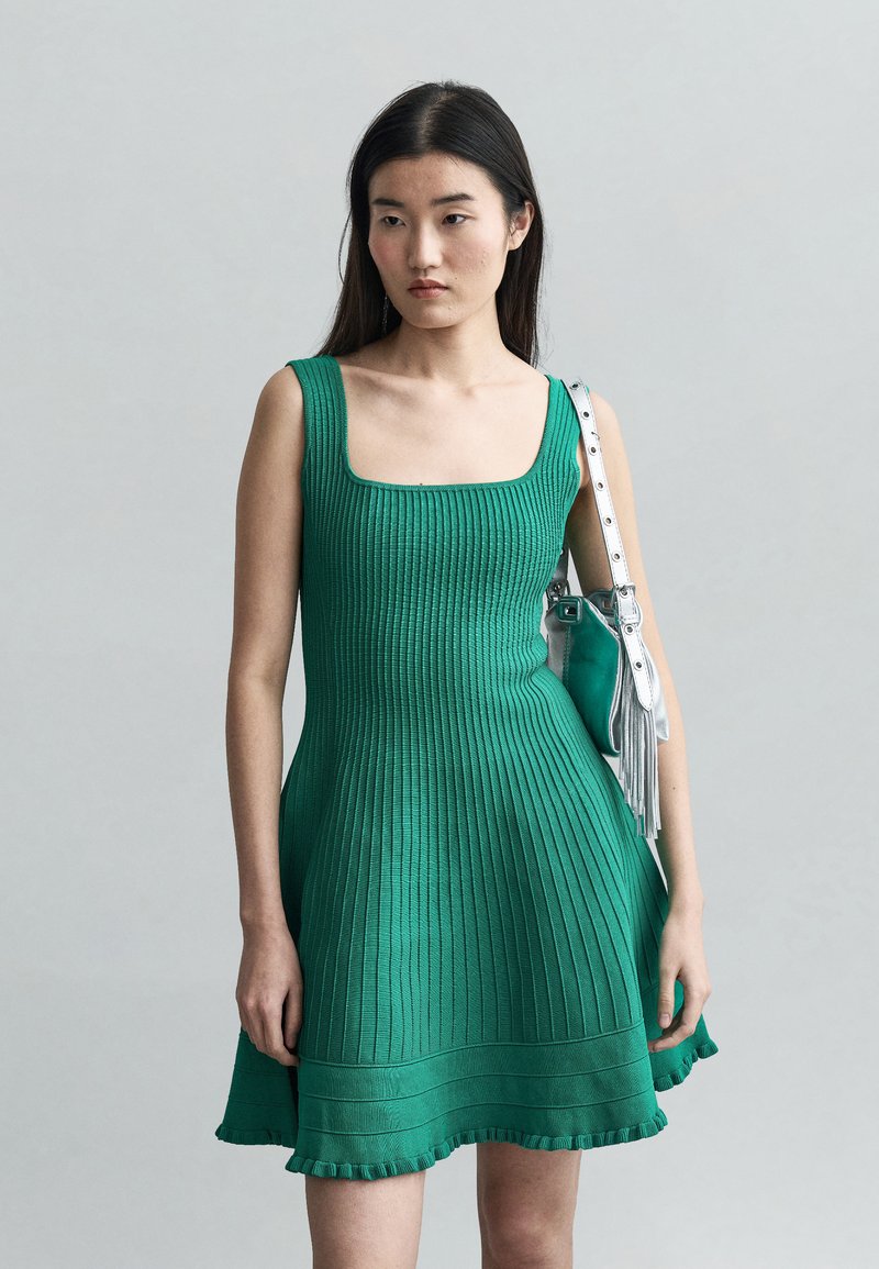 Jeune femme portant une robe sans manches verte en maille côtelée, tenant un sac à main argenté à franges sur un fond gris clair.