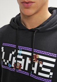 Sweat à capuche noir avec un motif ludique comprenant des personnages pixelisés, le mot "VANS" en blanc gras, et une bordure violette avec un motif d'échelle.