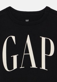 Μαύρο βαμβακερό μπλουζάκι με στρογγυλή λαιμόκοψη, με μεγάλο, έντονο κρεμ χρώματος κείμενο "GAP" στο στήθος. Ομαλή υφή υφάσματος.