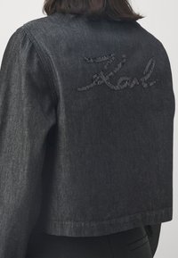 Giacca di denim nera con design cropped, caratterizzata da un logo ricamato testurizzato sulla schiena. Il tessuto ha una leggera lucentezza.