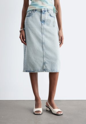 Kvinde iført lyseblå denim midi nederdel, hvide åbne blokhælede sandaler og lyseblå top mod en ensfarvet baggrund.