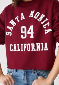 Persona che indossa un felpa bordeaux con testo bianco "Santa Monica 94 California" leggermente infilata in un paio di jeans blu.