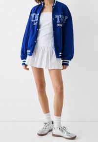 En person som bär en blå varsityjacka över en vit plisserad kjol och topp, i kombination med vita strumpor och silverfärgade sneakers.