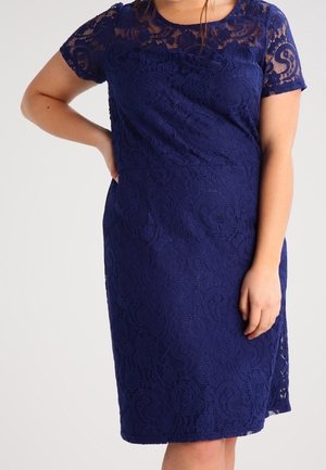 Kobieta w granatowej koronkowej sukience do kolan z krótkimi rękawami, jedna ręka na biodrze, druga swobodnie wzdłuż ciała.