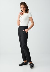 Femme portant un polo blanc à manches courtes, pantalon noir sur mesure, mocassins noirs, debout avec une main dans la poche.