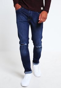 Mörkblå denimjeans med slitna knän, slim fit och upprullade ben. Kombineras med vita sneakers för en avslappnad look.