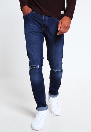 Vaqueros de mezclilla azul oscuro con rodillas desgastadas, ajuste slim y dobladillos enrollados. Combinados con zapatillas blancas para un look casual.
