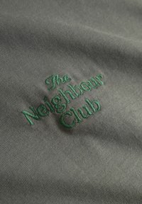 Texte brodé vert « The Neighbour Club » sur tissu gris, montrant la texture du tissu et les détails de la couture.
