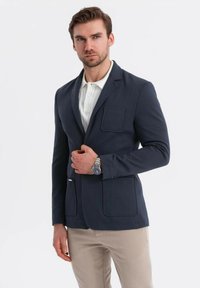 Man i marinblå kavaj över vit skjorta och beige byxor, justerar kavajen med en hand och bär en silverfärgad klocka.
