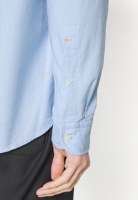 Lichtblauwe hem met lange mouwen van katoen. Beschikt over een knoopmanchet met witte knopen, een zijsplit en subtiele oranje stiksel details.