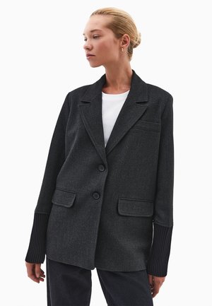 Donna con capelli biondi raccolti in un bun, che indossa un blazer oversize grigio scuro con maniche a costine nere e una camicia bianca sotto.