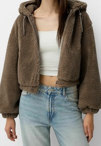 Bruine, korte fleecejas met capuchon, ritssluiting aan de voorkant en trekkoorden. Gecombineerd met een lichtblauwe, hoog taille denim jeans.