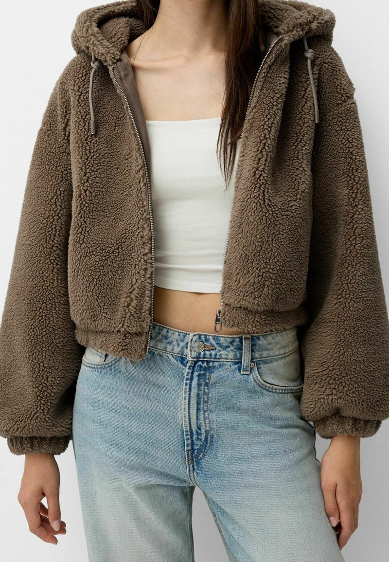 Bruine, korte fleecejas met capuchon, ritssluiting aan de voorkant en trekkoorden. Gecombineerd met een lichtblauwe, hoog taille denim jeans.