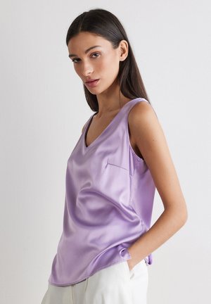 Kobieta z długimi ciemnymi włosami, ubrana w bezrękawnikowy lawendowy satynowy top z dekoltem w serek i białe spodnie, pozująca na jednolitym jasnym tle.