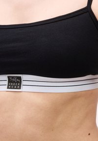 Μαύρο αθλητικό σουτιέν με λευκή ελαστική ταινία και μαύρες ρίγες, φορεμένο σε γυμνό κορμό, με εμφανή την ετικέτα της μάρκας "BJörn Borg UNDERWEAR" στην ταινία.