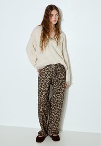 Młoda kobieta z długimi, falowanymi włosami, ubrana w beżowy, oversize'owy sweter, spodnie w panterkę oraz ciemne, puszyste kapcie, stojąca na tle białej ściany.