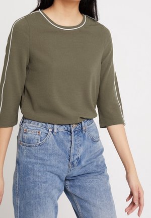 Kobieta w oliwkowozielonym długim rękawie z białym wykończeniem na rękawach i dekolcie, w połączeniu z jasnoniebieskimi, wysokim stanem jeansami.