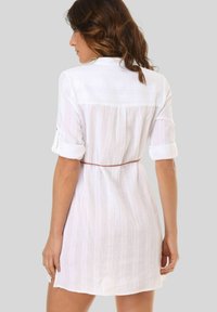 Vestido blanco de lino con un acabado texturizado, mangas arremangadas y cintura ajustada con lazo. Presenta un corte recto y un panel trasero en contraste.