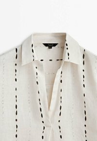 Camicia bianca di lino con un motivo astratto nero, dotata di colletto a punta e una parte di bottoni. Texture morbida e design leggero.
