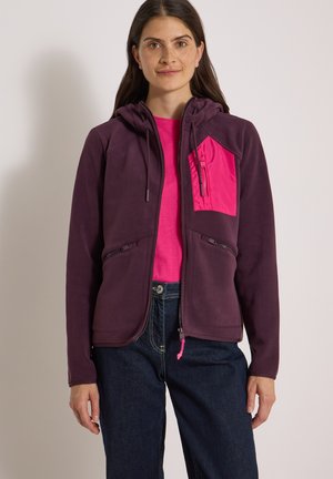Femme portant un sweat à capuche zippé violet foncé avec une poche poitrine rose vif sur un t-shirt rose et un jean bleu foncé, debout devant un fond uni.