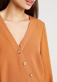 Blusa arancione bruciato con scollatura a V profonda, maniche lunghe e tre bottoni rotondi e decorati lungo la parte anteriore. Tessuto morbido.